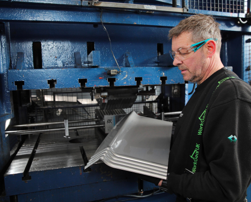 A man with glasses is holding a sheet of metal, indicating his involvement in a project or craft activity