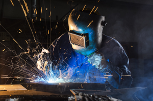 A worker welds metal in a factory, wearing protective gear and a welding torch