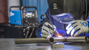 A welder in a helmet is actively welding a pipe, surrounded by tools and equipment in a workshop environment. 