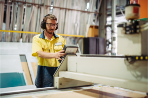 A man wearing a yellow shirt and headphones operates a machine in a workshop setting